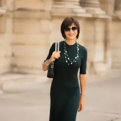 Une femme élégante porte une robe midi vert foncé, un collier de perles et des lunettes de soleil. C'est quoi une robe midi ? C'est une robe qui arrive à mi-mollet.
