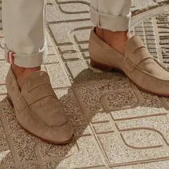 Un homme porte un mocassin à la mode en daim beige, avec un pantalon retroussé, sur un trottoir en dalles.