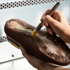 Un cordonnier applique de la colle sur la semelle d'une chaussure marron. Oui, on peut ressemeler des baskets, c'est un art !