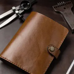 Portefeuille en cuir brun, entouré d'outils de **tannage du cuir** : compas, poinçon et scalpel.