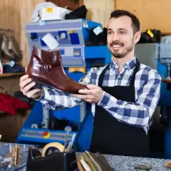 Artisan souriant tenant une botte en cuir marron, prêt pour la réparation de chaussure. L'atelier est bien équipé.