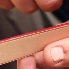 Main d'artisan appliquant une teinture de tranche cuir rouge vif sur une pièce de cuir clair.