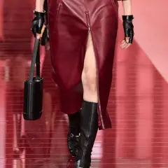 Mannequin portant une jupe courte en cuir bordeaux fendue et des bottines noires. Sac cylindrique noir et gants en cuir.