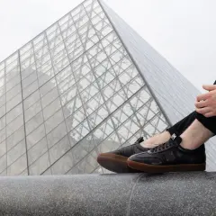 Un homme porte une tenue avec basket noir, assis devant la pyramide du Louvre.