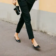 Une femme élégante marche dans la rue, portant un pantalon noir et des chaussures ballerines noires. Elle tient un petit sac noir.