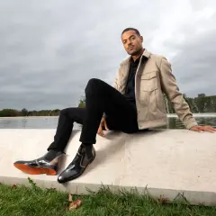 Un homme élégant montre comment porter des chelsea boots homme avec un jean noir et une veste beige, près d'une fontaine.