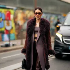 Une femme porte un manteau long en fausse fourrure prune, un exemple de types de manteaux femme tendance. Elle tient un sac noir.