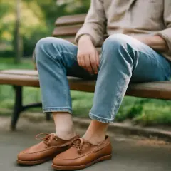 Un homme est assis sur un banc, portant une chemise beige, un jean et une paire de chaussure bateau chaussette en cuir marron.