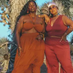 Deux femmes aux courbes généreuses posent dans le désert. L'une porte une robe longue orange, l'autre un ensemble pantalon et top rouge. Des tenues parfaites pour savoir comment s'habiller quand on a des grosses cuisses.