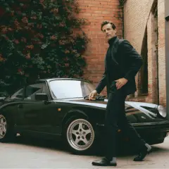 Un homme élégant en costume et bottines noires pose près d'une Porsche verte.