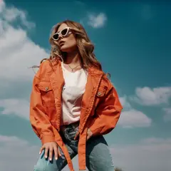 Une jeune femme porte une tenue mi saison femme stylée : veste orange, t-shirt blanc et jean. Elle est dehors, sous un ciel bleu nuageux.