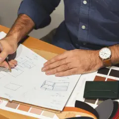 Un designer esquisse des modèles de maroquinerie, entouré d'échantillons de cuir aux teintes variées, évoquant le tannage au chrome.