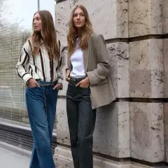 Deux femmes posent devant un mur en pierre. L'une porte un jean tapered femme, une chemise rayée et une ceinture chaîne. L'autre, un blazer et un jean.