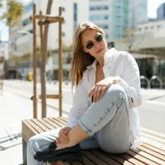 Une femme assise sur un banc, portant un chemisier blanc, un jean décontracté et des mocassins noirs. Son outfit simple et chic est parfait pour une journée en ville.
