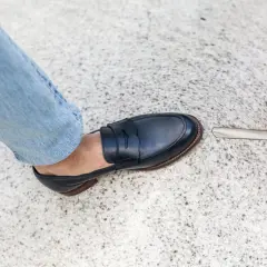 Un pied en jean bleu porte un mocassin bleu marine. La semelle extérieure de la chaussure, signée par un cordonnier, est visible.