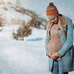 Une femme enceinte, bien emmitouflée dans un manteau et un bonnet coloré, pose dans un paysage enneigé. Elle montre comment s'habiller enceinte en hiver avec style et confort.