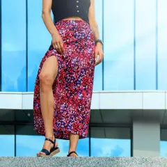 Une femme en jupe longue à fleurs et escarpins bleus, posant devant un bâtiment moderne aux reflets bleus.