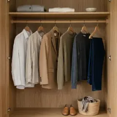 Garde-robe en bois clair, organisée avec des vêtements de couleur sobre : chemises, veste, pulls et jean. Chaussures et panier en osier au sol.