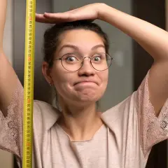 Une femme trapue, lunettes rondes, mesure sa taille avec un mètre ruban jaune. Elle a une expression amusée.