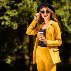 Une femme en costume jaune vif, chapeau et lunettes de soleil, tenant un café. Parfait pour savoir comment s'habiller quand il fait 14 degrés.