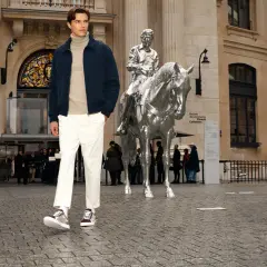 Un homme porte une chaussure avec un chino blanc, un pull à col roulé beige et une veste bleu marine devant une sculpture de cheval argenté.