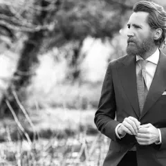 Un homme barbu en costume sombre, chemise blanche et cravate grise, ajuste sa veste. Sa tenue avec short blanc homme est élégante.
