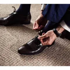 Un homme en costume bleu marine attache ses chaussures vernies marron. Quel chaussure avec un costume pour un look impeccable.