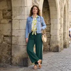 Une femme pose devant des arches en pierre. Elle porte une veste en jean, un chemisier à motifs, un pantalon vert et des sandales. Idéal pour savoir comment s'habiller quand il fait 12 degrés femme.
