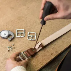 Artisan travaillant sur une ceinture en cuir, utilisant un tournevis pour fixer une boucle. Outils de mesure et boucles supplémentaires à proximité.