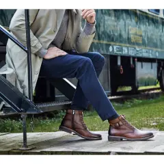 Un homme élégant, assis près d'un train, porte des bottines marron. Une inspiration pour savoir quelles chaussures porter avec un jean homme.