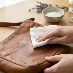 Main d'une personne en train d'enlever une tache sur un sac en cuir clair avec un chiffon blanc. Vinaigre, citron et lavande en arrière-plan.