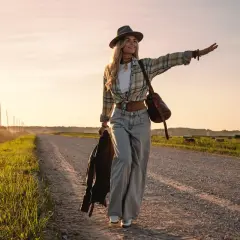Une femme au style bohème, portant un jean large, fait de l'auto-stop au coucher du soleil. Elle incarne la liberté et les différents types de jean femme.