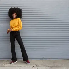 Une jeune femme souriante pose devant un mur gris, portant un haut jaune et un jean flare. Quelles chaussures avec jean flare ? Des baskets bordeaux complètent ce look décontracté.