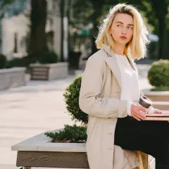 Une femme blonde, assise dehors avec un livre et un café, porte un trench beige et un haut blanc. Parfait pour savoir comment s'habiller quand il fait 15 degrés.