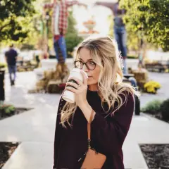 Une femme blonde avec des lunettes boit un café. Elle porte un pull bordeaux. Elle se demande comment dois je m habiller demain.
