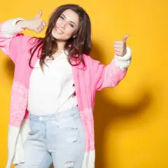 Une femme souriante, petite et ronde, montre comment s'habiller avec un gilet rose et un jean. Elle fait un signe de pouce levé.