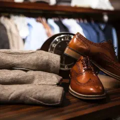 Chaussures en cuir marron élégantes, prêtes à être assouplies. Un pull plié et un manteau sont visibles en arrière-plan.