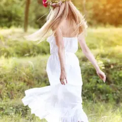 Une jeune femme aux cheveux blonds, portant une couronne de fleurs et une robe blanche légère, incarne l'esprit du printemps. Elle danse dans un champ verdoyant, une vision parfaite pour savoir comment s'habiller au printemps.