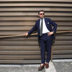 Un homme élégant porte un costume bleu marine, une chemise blanche et une cravate à motifs. Ses chaussures marron complètent parfaitement sa tenue.