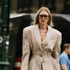 Une femme porte une tenue avec un blazer blanc chic, des lunettes de soleil oversize et un top en dentelle. Elle déambule dans la rue.