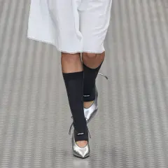 Une mannequin défile avec une tenue blanche et des chaussettes hautes grises, rehaussée par des chaussures argentées pointues.