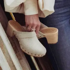 Une femme tient un sabot femme en toile beige, orné de clous, avec un talon en bois. Elle porte un jean bleu.