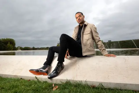 Un homme élégant montre comment porter des chelsea boots homme avec un jean noir et une veste beige, près d'une fontaine.