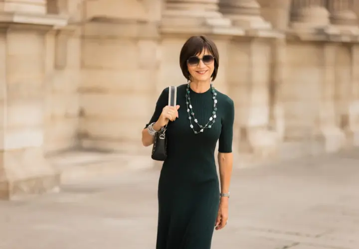 Une femme élégante porte une robe midi vert foncé, un collier de perles et des lunettes de soleil. C'est quoi une robe midi ? C'est une robe qui arrive à mi-mollet.