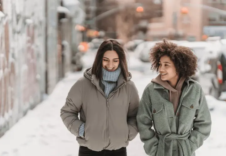 Deux amies profitent d'une promenade hivernale, bien emmitouflées dans leur vêtement contre le froid. Le top 7 des tenues chaudes pour affronter la neige.