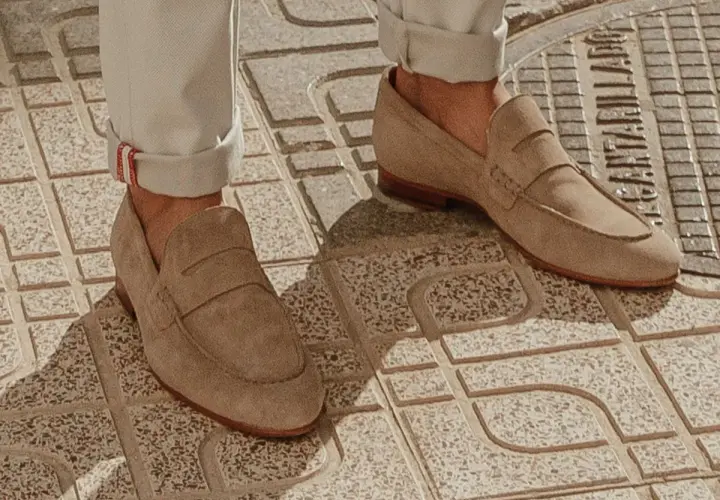 Un homme porte un mocassin à la mode en daim beige, avec un pantalon retroussé, sur un trottoir en dalles.