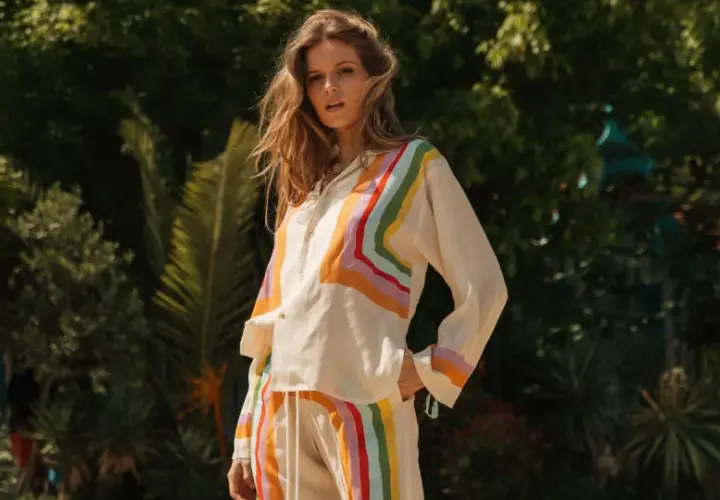 Une femme pose dans un jardin luxuriant, vêtue d'un ensemble aux rayures arc-en-ciel, évoquant la couleur de l'été.