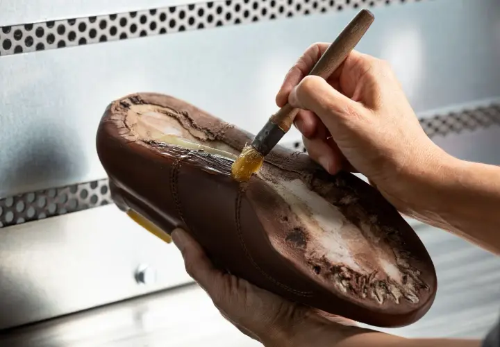 Un cordonnier applique de la colle sur la semelle d'une chaussure marron. Oui, on peut ressemeler des baskets, c'est un art !