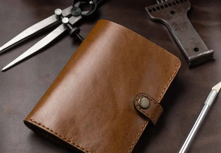 Portefeuille en cuir brun, entouré d'outils de **tannage du cuir** : compas, poinçon et scalpel.