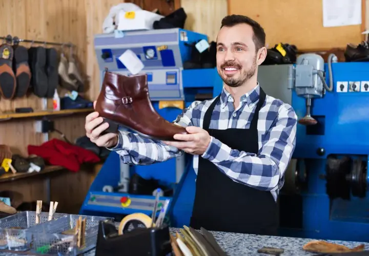 Artisan souriant tenant une botte en cuir marron, prêt pour la réparation de chaussure. L'atelier est bien équipé.